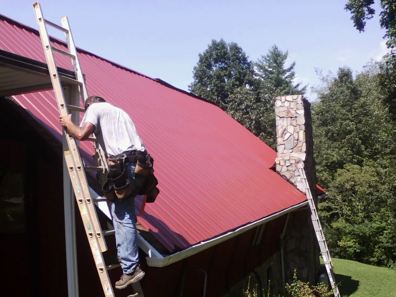 Roofer on a ladder beside a red metal roof and stone chimney — Soffit & Fascia Repair by Roofing Brothers in Walnut